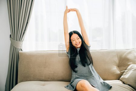 Foto de Mujer sonriente en el sofá levanta los brazos se estira disfrutando de la relajación después de sentarse. Estilo de vida de un freelancer feliz en la sala de estar de lujo. Bienestar alegría y celebración despreocupada en casa. - Imagen libre de derechos