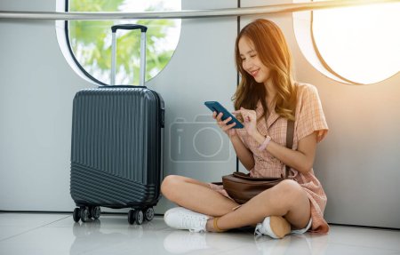 Photo for Young woman sitting next to her suitcase and backpack at the airport terminal, using her phone to book her next travel destination. travel in vacation summer - Royalty Free Image