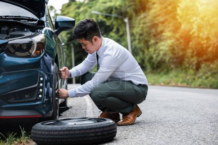 Photo for Asian businessman with lug wrench changing car wheel on the highway. Professional car mechanic providing road assistance. Vehicle tyre replacement concept. - Royalty Free Image