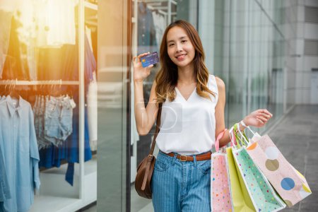 Foto de Una joven con una sonrisa sostiene bolsas de compras y una tarjeta de crédito frente a un escaparate. Ella está dispuesta a gastar dinero y aprovechar los últimos descuentos y ofertas - Imagen libre de derechos