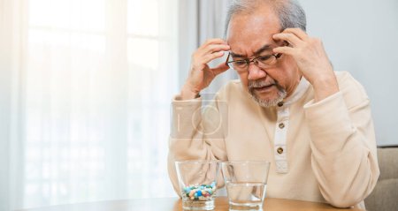 Photo for Asian senior old man grey haired sad sitting in chair hand hold head with many medicines and pills for taking, elderly man sick have headache looking pill glass on at home, Elderly healthcare - Royalty Free Image