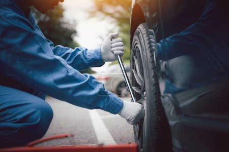 Foto de Mecánico de automóviles calificado en uniforme de trabajo azul, en cuclillas junto a un vehículo en un taller de reparación de automóviles, la fijación de un neumático con un enfoque en las manos. - Imagen libre de derechos