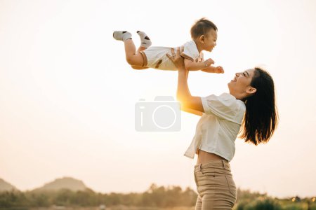 Foto de Una mujer sostiene a su bebé recién nacido en alto, ya que disfrutan de un momento de naturaleza en el parque al atardecer. El pequeño mira al cielo con asombro, mientras la orgullosa mamá captura el precioso momento de la vida - Imagen libre de derechos