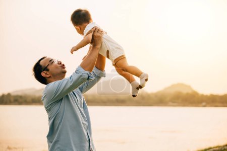 Téléchargez les photos : Un père heureux et son fils tout-petit partagent un moment ludique de liberté et de joie dans le parc, le jetant dans les airs par une journée d'été ensoleillée. Amour de famille et bonheur capturés dans une photographie - en image libre de droit
