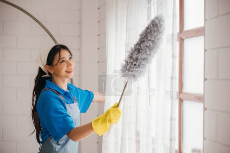 Photo for Smiling womans portrait during window cleaning with a duster. Standing she enjoys routine housework ensuring hygiene. Modern cleaning occupation for a clean and sparkling home. whisk - Royalty Free Image