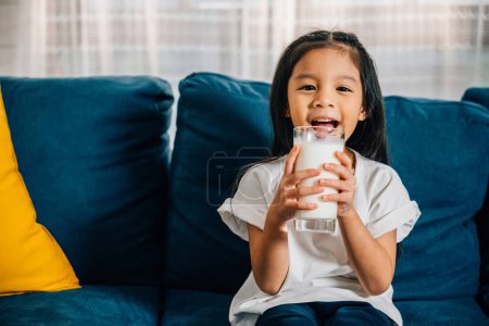 Téléchargez les photos : Dans un appartement confortable, une fille asiatique joyeuse boit du lait dans un verre tout en étant assise sur le canapé. Cette image chaleureuse met l'accent sur les soins de santé quotidiens et l'innocence joyeuse de l'enfance. - en image libre de droit