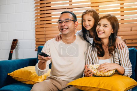 Téléchargez les photos : Une jeune famille souriante avec du pop-corn se réunit dans le salon en regardant la télévision ensemble. Ils profitent de moments francs de liaison et de détente pendant leur séjour de qualité à la maison. - en image libre de droit