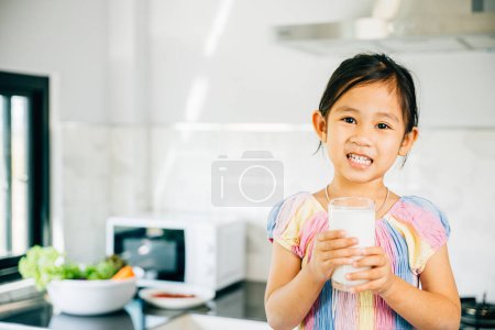 Téléchargez les photos : Adorable préscolaire asiatique tient tasse de lait dans la cuisine. Jolie fille est assise souriant avec joie profiter de la boisson. Portrait de fille heureuse à la maison savourant du bonheur liquide riche en calcium me donne. - en image libre de droit
