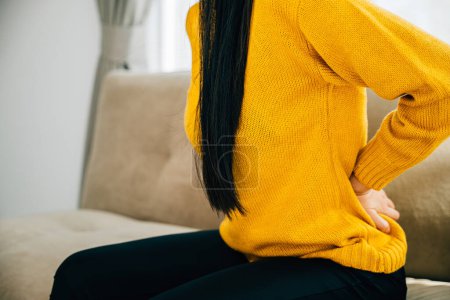 Photo for Portraying backache, Asian woman on sofa suffers from unbearable lower back pain. Illustrating chronic back pain discomfort and necessity of medical care for back problems. health care concept - Royalty Free Image