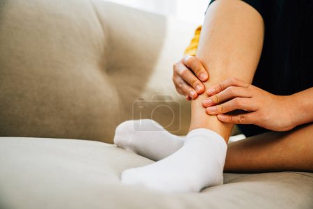 Photo for Illustrating health care concept, woman sits on sofa holding her painful ankle. Emphasizing varicose vein prevention leg recovery and pain management for the patient. medical - Royalty Free Image