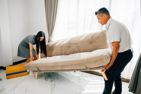 Téléchargez les photos : Un couple asiatique sourit en déplaçant un canapé dans leur nouvel appartement, mettant en valeur le travail d'équipe et l'excitation liés à la réinstallation. Parfait pour illustrer le remplacement des meubles et s'installer dans une maison confortable. - en image libre de droit
