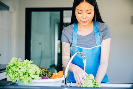 Téléchargez les photos : Belle femme savoure la cuisine et le nettoyage des légumes frais dans l'évier de la cuisine pour une salade saine. Mettre l'accent sur la propreté et le plaisir de la nourriture végétarienne pendant les vacances d'été. - en image libre de droit