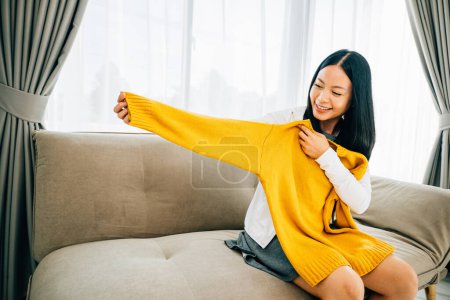 Photo for Smiling Asian woman unwraps a shirt from a box excited at home. Delighted customer reveals online package exhibiting new clothes. Delivery and online shopping concept. - Royalty Free Image