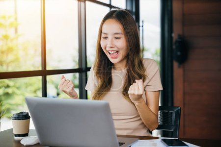 Photo for Excited female feeling winner rejoicing online win got new job on laptop computer at home, happy Asian business young woman sitting at cafe desk raising hands up and celebrating success - Royalty Free Image
