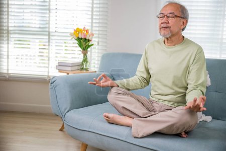 Photo for Lifestyle senior man sitting on sofa in living room holding hands in mudra practicing home yoga in lotus pose, Elderly relaxed man do physical exercises following healthy on stress free weekend - Royalty Free Image