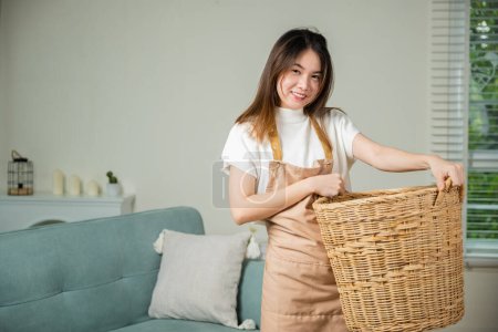 Téléchargez les photos : Femme au foyer asiatique tenant panier avec tas de vêtements différents sur le canapé dans le salon, belle femme de ménage faisant des travaux ménagers tient panier en bois de vêtements propres à laver à la maison - en image libre de droit