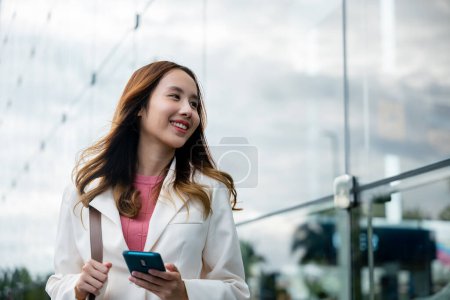 Téléchargez les photos : Asian businesswoman working with mobile phone standing against street front building near office, Portrait successful business woman smiling holding smartphone use application chat online in morning - en image libre de droit