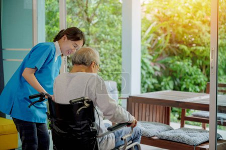 Téléchargez les photos : Jeune infirmière asiatique soutient homme âgé assis en fauteuil roulant marchant à regarder par la fenêtre sur le jardin et respirer l'air frais, centre de retraite, homme âgé à la maison, concept de soins de santé - en image libre de droit