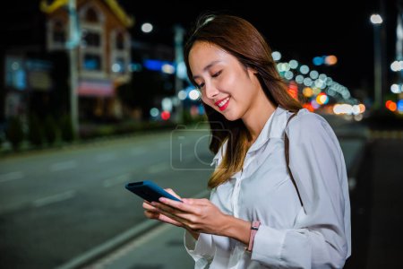 Foto de Una mujer se para en la acera por la noche, sosteniendo su teléfono celular mientras espera el transporte, con una vibrante combinación de colores naranja y verde azulado. - Imagen libre de derechos