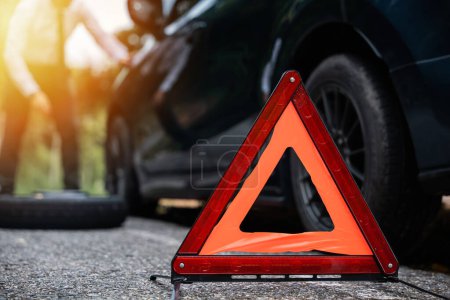 Photo for Businessman stranded on the road with a broken car and a warning triangle sign. Repairing the car wheel while waiting for roadside assistance. Concept of transportation problems and emergency support. - Royalty Free Image