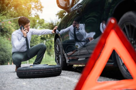 Photo for Broken down car with a red triangle warning sign on the road. Businessman waiting for assistance while leaning on the car wheel. Concept of car breakdown and roadside rescue service. - Royalty Free Image