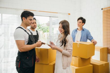 Photo for Couple inspects checklist before signing moving papers. Worker carries a box. Professional movers ensure seamless home relocation. Teamwork and efficiency displayed. Movind Day to New home - Royalty Free Image