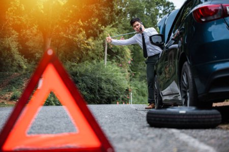 Photo for Asian businessman standing by broken down car, calling for help and showing thumbs up. Warning triangle behind car and man waiting for roadside assistance. Precautions and safety concept. - Royalty Free Image