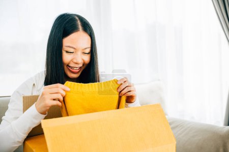 Photo for Overjoyed woman unpacks online delivery holding a shirt on the couch. Her smile reflects happiness and satisfaction with the received dress. Satisfied buyer concept. - Royalty Free Image