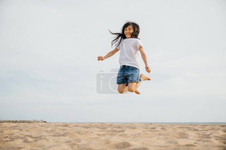 Téléchargez les photos : Capturer l'essence de l'enfance une petite fille saute sur la plage sous le soleil des Caraïbes. Vitalité de mouvement ludique et portrait de joie et de bonheur - en image libre de droit