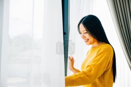Photo for A young woman opens the curtains in the early morning smiles at the pleasant view feeling refreshed and joyful at home. Embracing morning happiness relaxation and a cheerful start. - Royalty Free Image