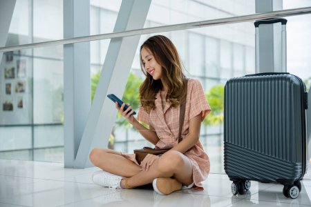 Téléchargez les photos : Femme assise sur le sol avec sa valise et parcourant les médias sociaux sur son téléphone dans le terminal de l'aéroport. Voyage touristique concept de voyage - en image libre de droit