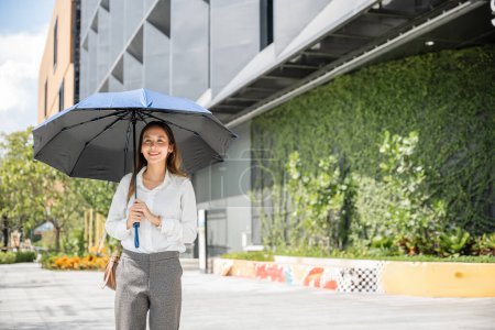 Foto de Caminando a la oficina en un día abrasador, una joven empresaria sostiene un paraguas para protegerse del sol caliente. Su determinación y sudor resaltan su compromiso con el éxito. - Imagen libre de derechos