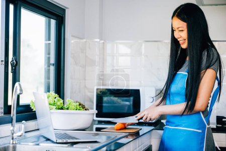 Photo for A woman happily cooks in the kitchen using a laptop for cooking videos or tutorials. Demonstrating the joy of technology assisting in culinary exploration. - Royalty Free Image