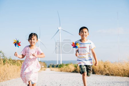 Foto de Hermano y hermana dirigidos por molinos de viento que sostienen molinos. Niños alegría cerca de las turbinas representa un futuro de energía limpia. Los niños sonrientes, despreocupados y felices, se comprometen con la tecnología sostenible. - Imagen libre de derechos
