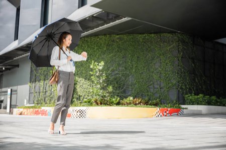 Foto de Caminando a la oficina bajo un clima caluroso, una mujer de negocios sostiene un paraguas, verificando el tiempo durante una ajetreada jornada laboral. Su aspecto severo y profesional significa su compromiso con el éxito. - Imagen libre de derechos