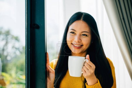 Foto de Una mujer sonriente sostiene una taza de café junto a la ventana disfrutando del amanecer matutino en casa. Reflejando la relajación de la alegría y la contemplación en las primeras horas. - Imagen libre de derechos