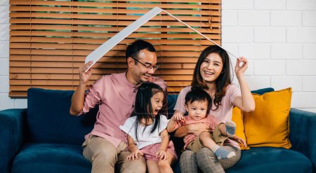 Photo for In their new house a smiling family sits on the sofa holding a cardboard roof mockup representing security and happiness during their home relocation. The key is family support and planning. - Royalty Free Image