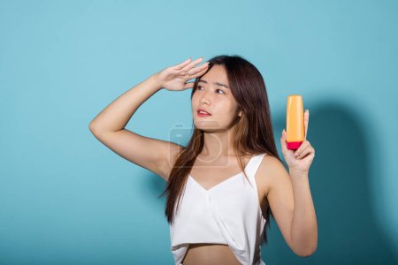 Photo for Beautiful Asian young woman holding body lotion on hand in studio shot isolated on blue background, Smiling brunette female hold cream sunblock bottle, tube of sunscreen, protect sun light and UV - Royalty Free Image