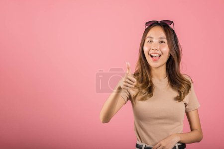 Foto de Retrato asiático hermosa joven mujer sonriendo ella de pie hizo dedo pulgares hacia arriba, Ok signo de acuerdo estudio tiro aislado sobre fondo rosa, tailandés femenino éxito como gesto de dedo con espacio de copia - Imagen libre de derechos