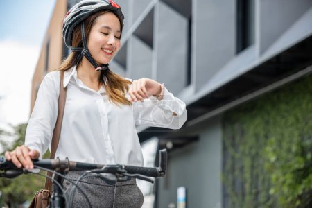 Téléchargez les photos : Le matin, une jeune femme d'affaires avec un vélo vérifie l'heure, incarnant le mode de vie d'un voyageur d'affaires. Son look professionnel et son casque reflètent sa profession d'entreprise. - en image libre de droit