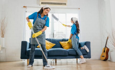 Foto de Dos mujeres sonrientes en uniforme en la sala de estar desordenada trabajando un equipo para el servicio de limpieza de la casa. Utilizando equipos profesionales cuidan el sofá de los muebles y la higiene. El trabajo doméstico es su rutina.. - Imagen libre de derechos