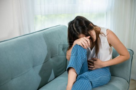 Photo for Asian young woman unhappy hands holding on stomach suffering from abdominal pain on sofa at home in living room, Sad female close eyes she painful stomachache, medical care and health concept - Royalty Free Image