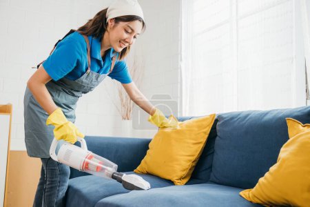 Photo for In modern setting Asian woman maid uses a vacuum machine cleaner to clean a sofa in a living room. Her dedication to hygiene and housework shines through as she focuses on furniture care. - Royalty Free Image