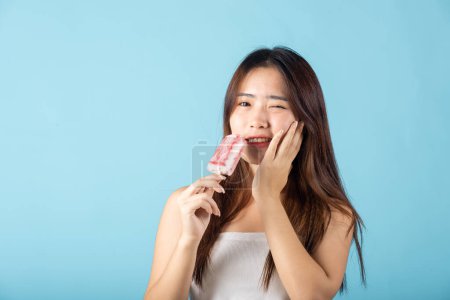 Photo for Portrait of Asian young woman with sensitive teeth after eating delicious ice cream wood stick mixed fruit flavor feeling painful uncomfortable, studio shot isolated on blue background, dental problem - Royalty Free Image
