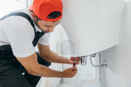 Photo for Skilled plumber using a wrench repairs a water pipe under a bathroom sink. His focus is on maintenance fixing leaks and improving pipelines for efficient plumbing service and cleaning. - Royalty Free Image
