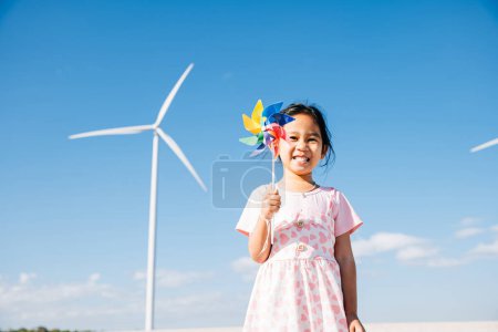 Foto de Niñas juguetonas alegría cerca de molinos de viento, corriendo con molinetes. Ilustra la educación en energía eólica promoviendo la electricidad limpia y la industria sostenible en un entorno pintoresco. - Imagen libre de derechos