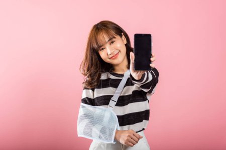Photo for Despite a broken arm, this woman smiles confidently, showing her smartphone screen. Coping with pain and managing medical expenses on a pink background. - Royalty Free Image