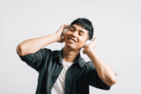 Téléchargez les photos : Heureux homme asiatique se livre à la joie d'écouter de la musique sur son smartphone, équipé d'écouteurs Bluetooth. Studio tourné isolé sur blanc, soulignant la beauté de la technologie moderne. - en image libre de droit