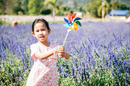 Photo for A cheerful little girl stands in a flower garden holding a toy pinwheel. The vibrant springtime evokes joy and flying pinwheels embodying childhood happiness and freedom in natures sunny embrace. - Royalty Free Image