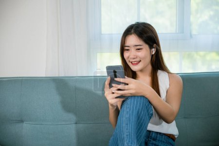 Téléchargez les photos : Joyeux jeune femme détendue et messagerie smartphone, Souriant asiatique femme assise sur le canapé en utilisant téléphone intelligent mobile pour faire du shopping en ligne dans le salon à la maison, Réseaux sociaux de communication - en image libre de droit